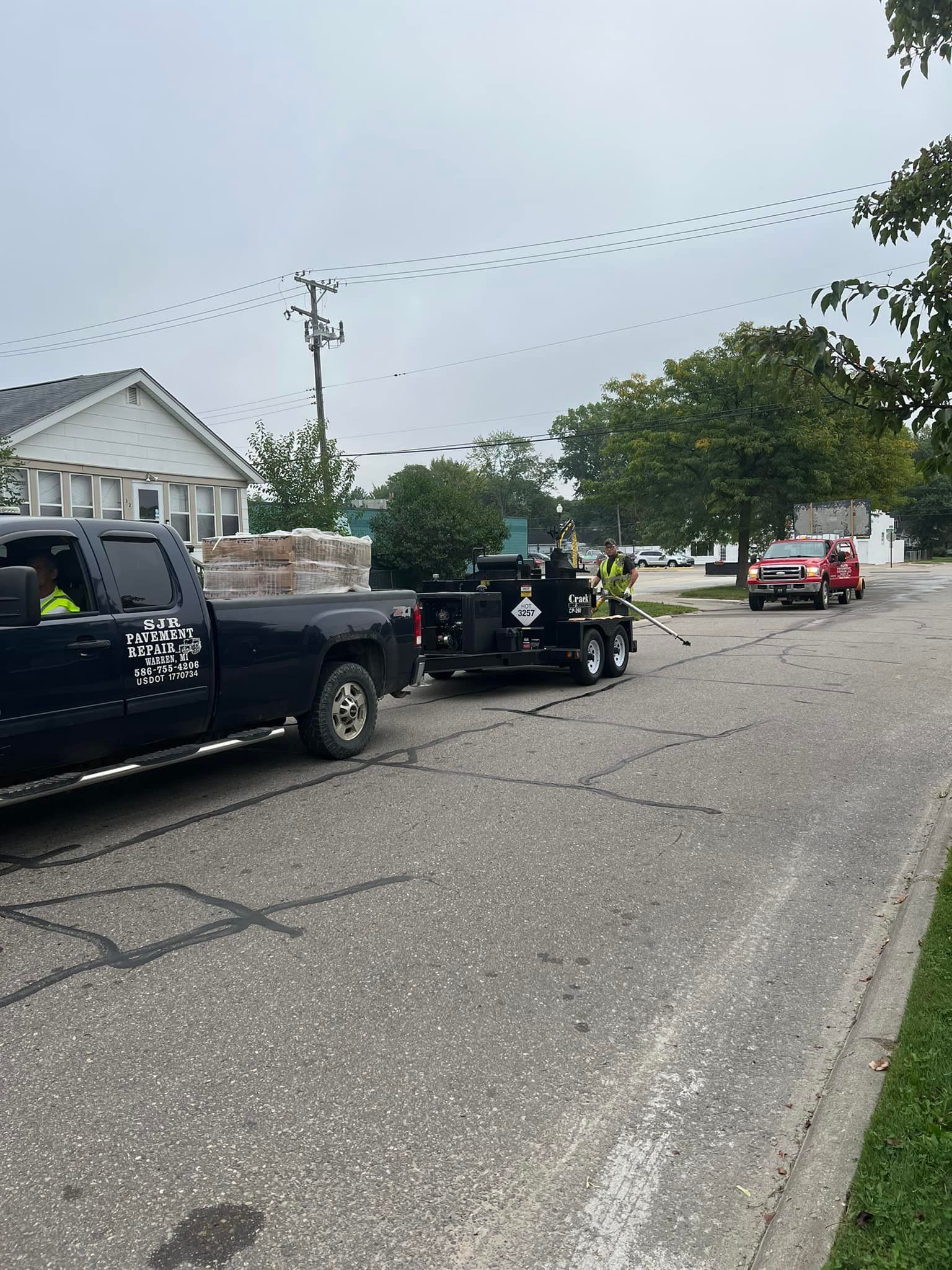 Black truck pulling a trailer with workers. Red truck in background on a street. Overcast sky.