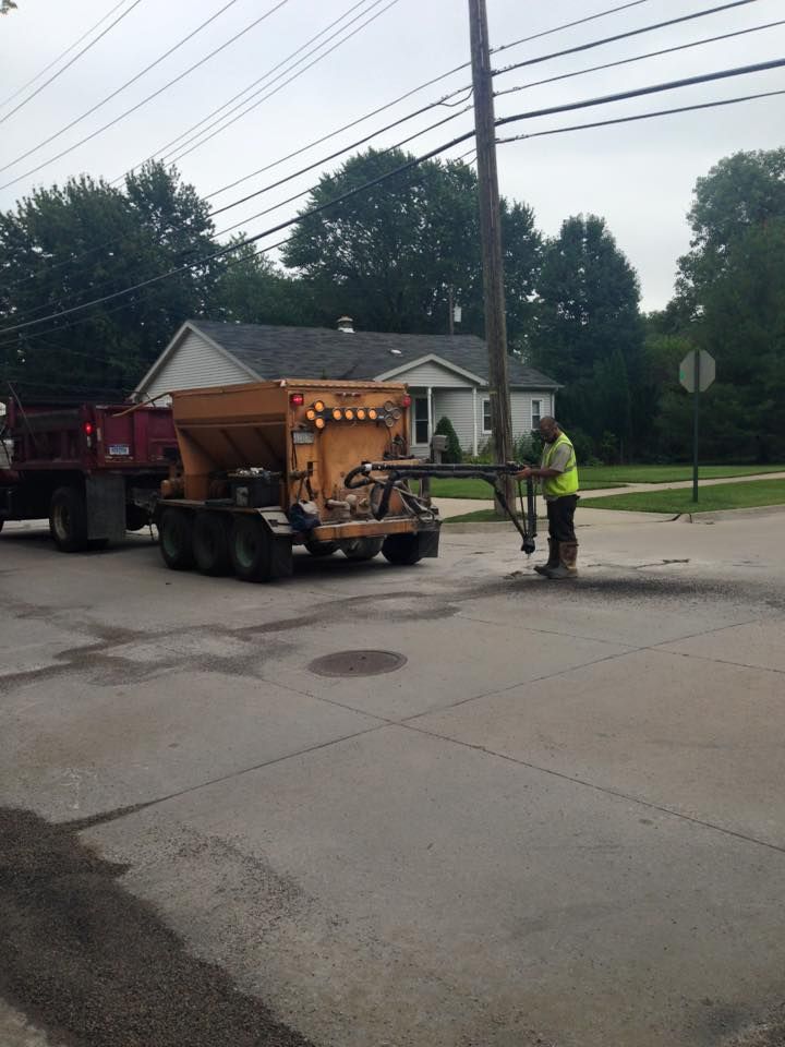 Worker in vest operating asphalt patching machine on a street, truck nearby, house in background.