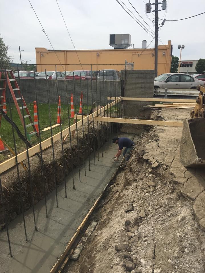 Construction worker in a trench with exposed rebar. Concrete formwork and orange cones are visible.