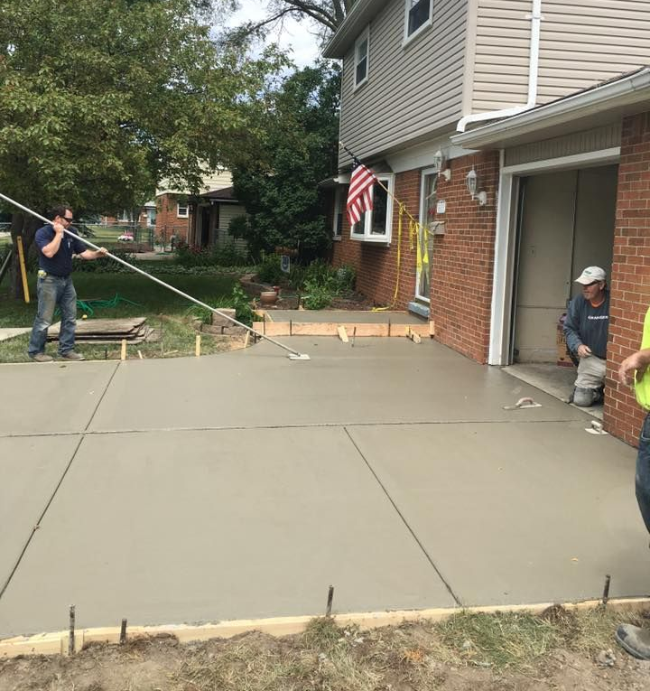 Workers finishing a concrete driveway next to a brick house. One holds a line, another kneels.