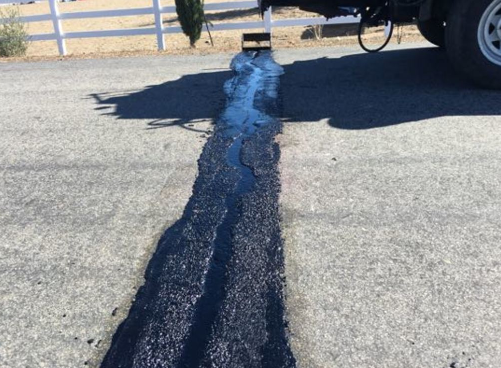 Black asphalt sealant fills a crack in a paved road; a truck is parked nearby.
