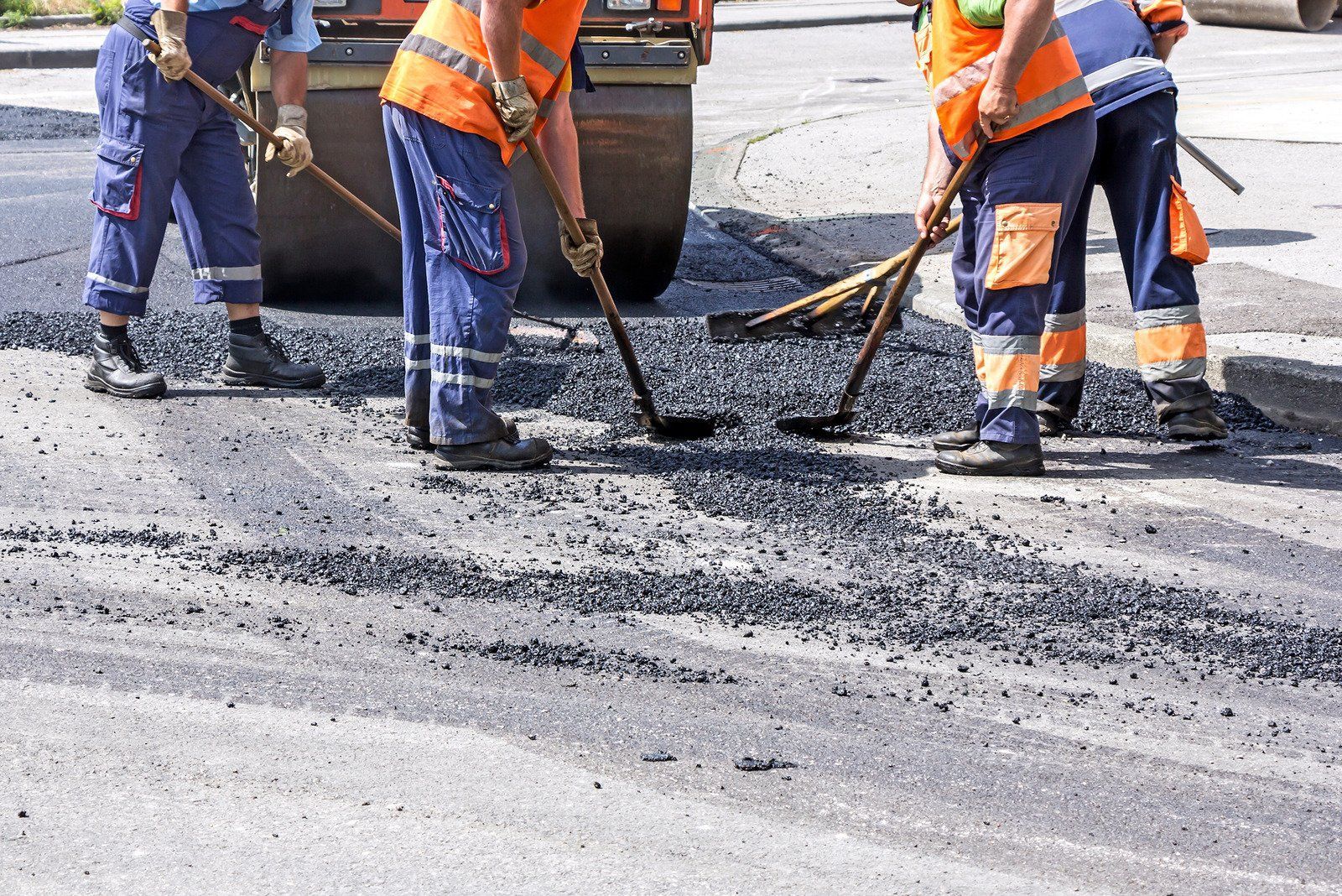 Road workers using rakes to spread asphalt, with a road roller in the background.