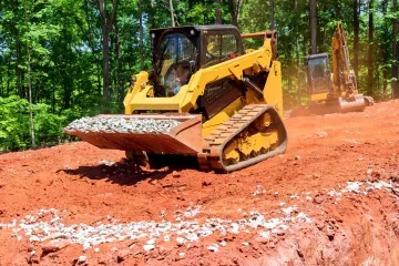 Yellow skid-steer loader on red dirt, moving gravel. Another machine in the background, trees in the background.