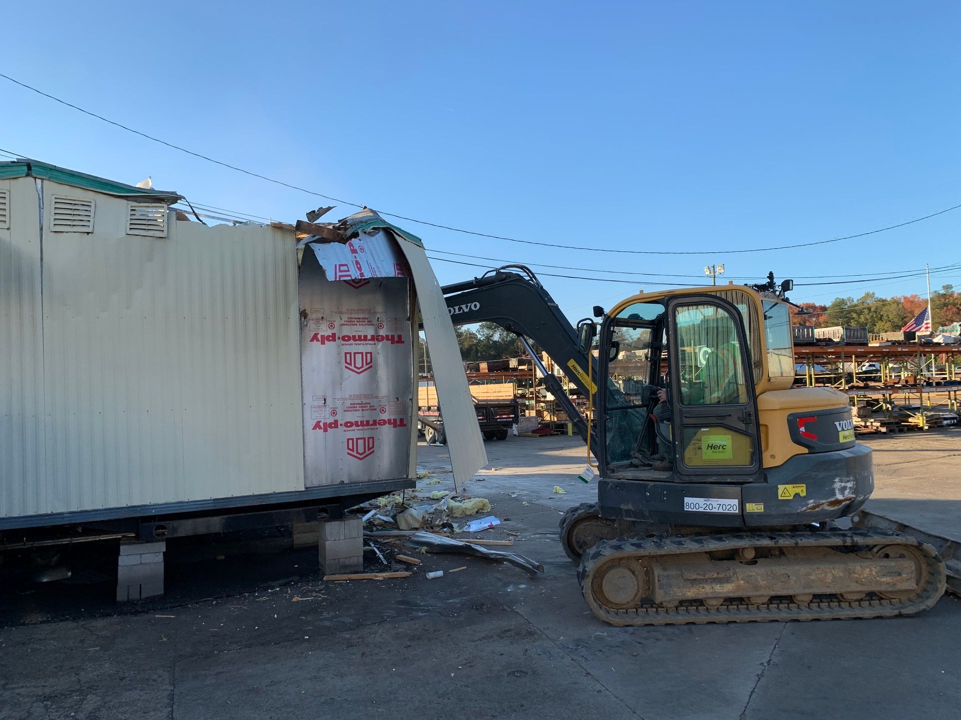 Yellow excavator demolishing a white mobile building outdoors on a sunny day.