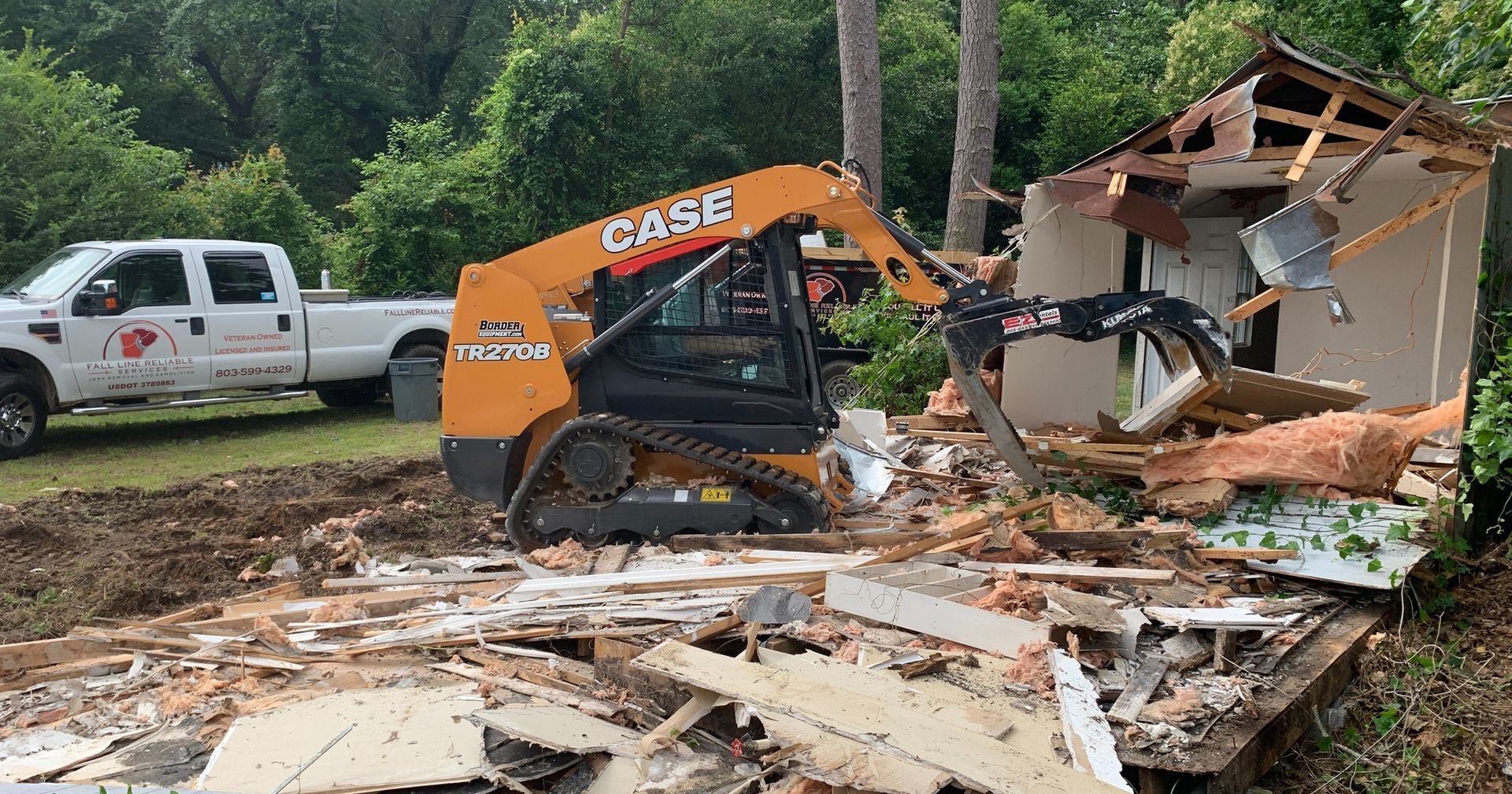 A Case skid steer demolishes a small building, debris scattered. Truck parked nearby. Green trees.