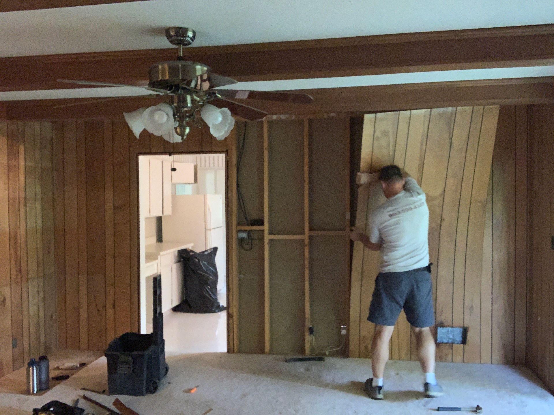 Man removing wood paneling from a wall in a room with exposed studs and an open doorway.