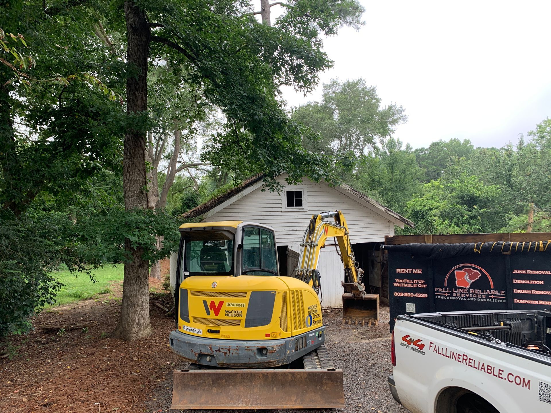 Yellow excavator in front of a white building; a truck and a dumpster are nearby.
