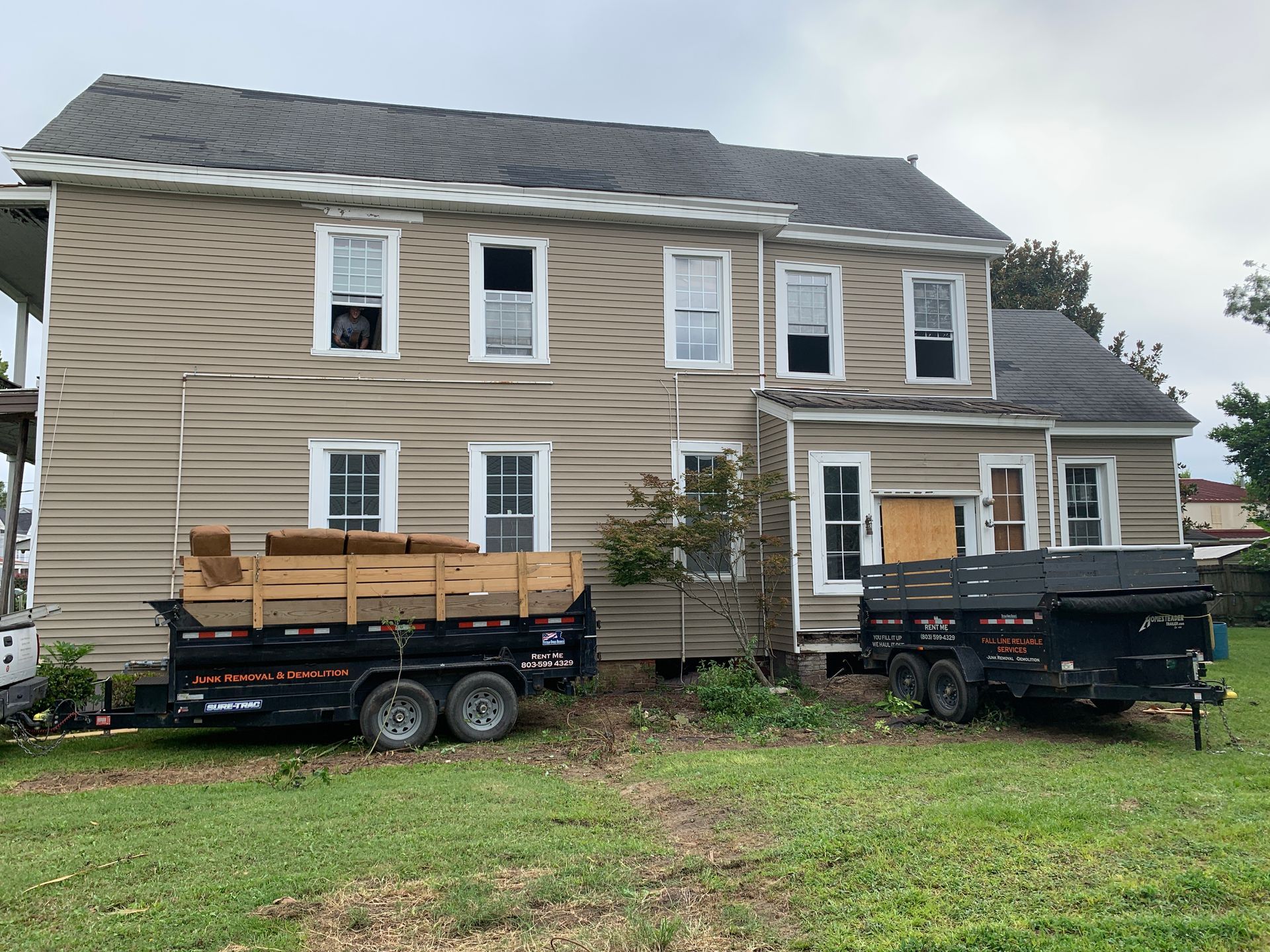 Two trailers next to a two-story tan house with boarded-up windows, cloudy sky above.