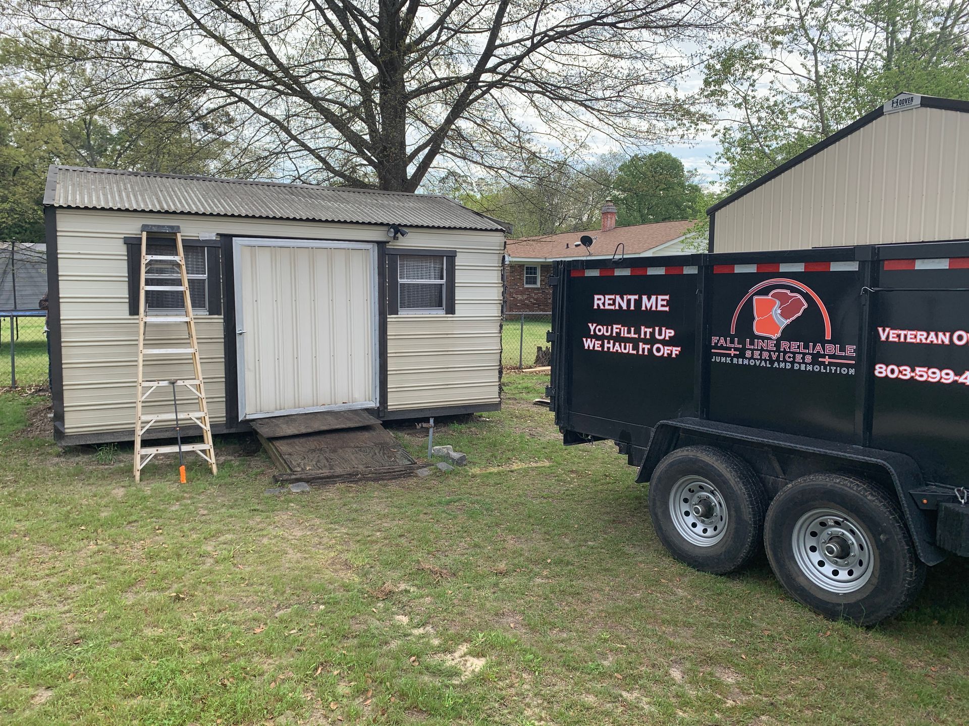 A shed with a ladder is next to a black dumpster trailer, in a grassy backyard.