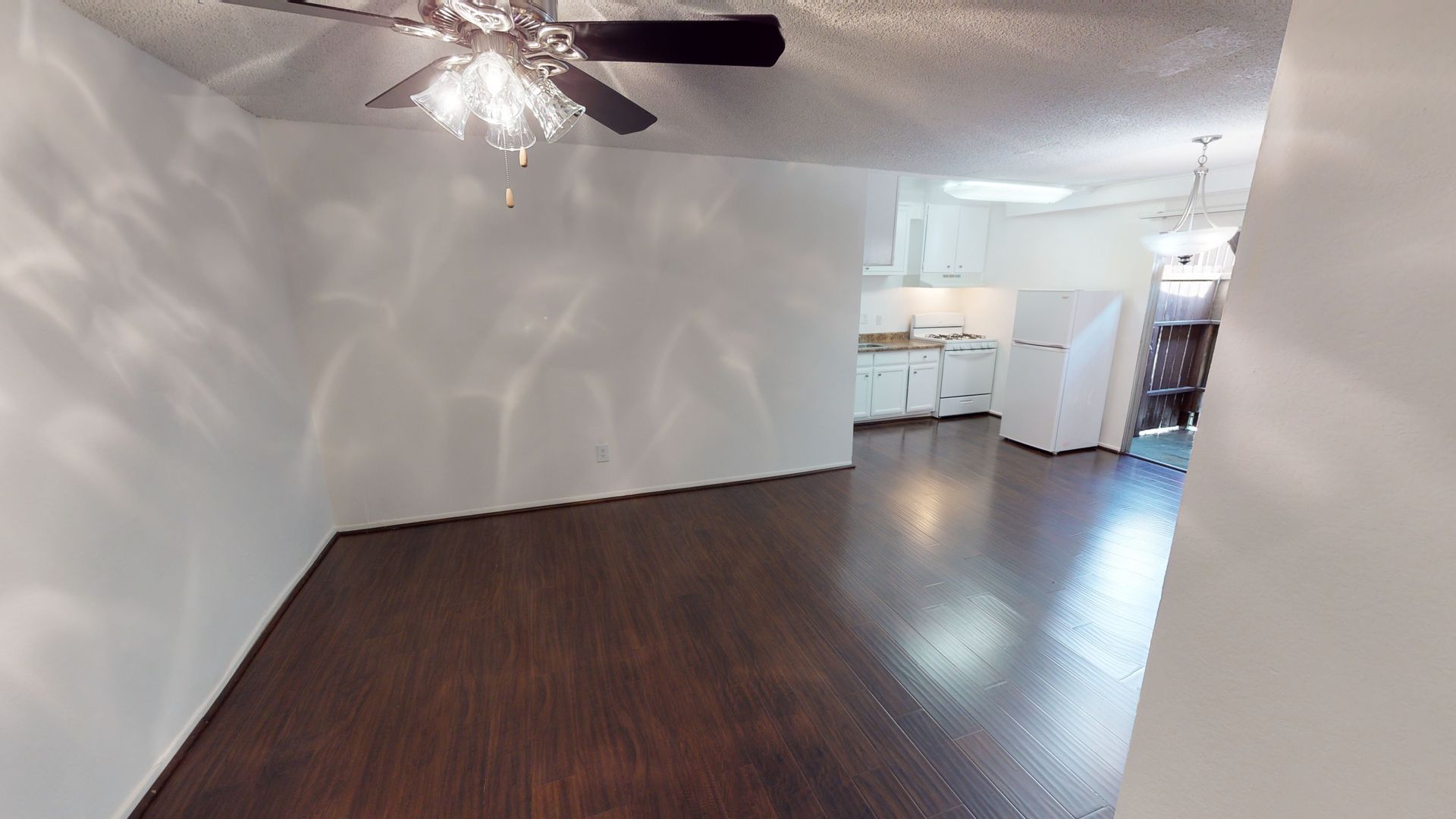 Empty living room with dark wood floor, white walls, and a ceiling fan. Kitchen visible in the background.