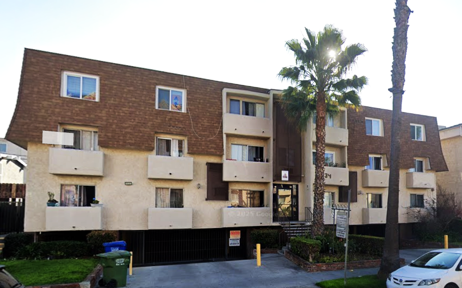 Three-story apartment building with tan facade, brown roof, and small balconies. Palm tree in front.