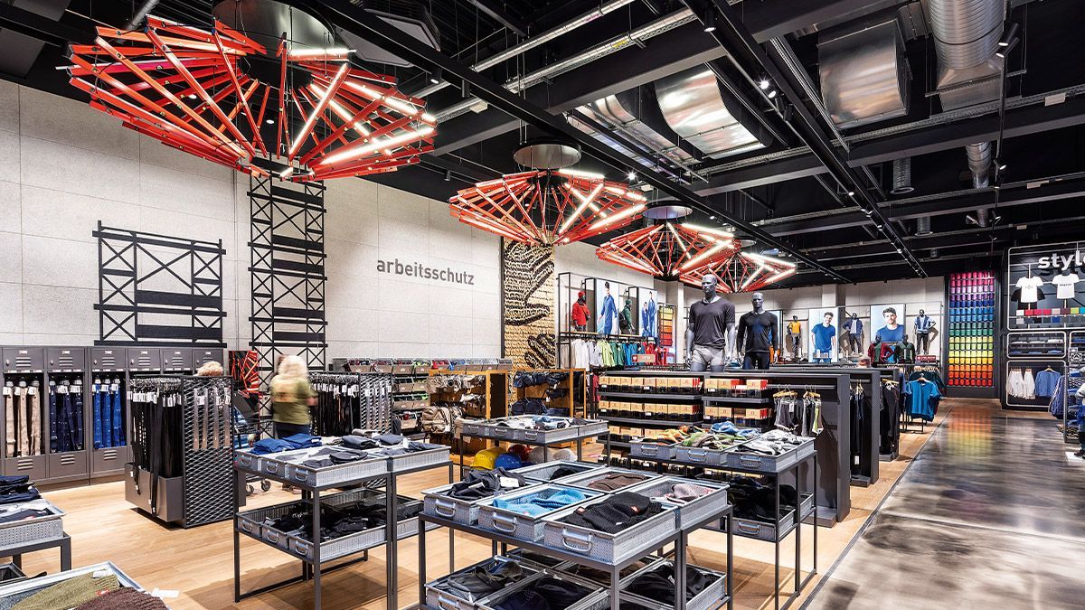 Inneneinrichtung eines Herrenbekleidungsgeschäfts mit rot beleuchteten geometrischen Deckenleuchten, Kleiderständern und Auslagen.