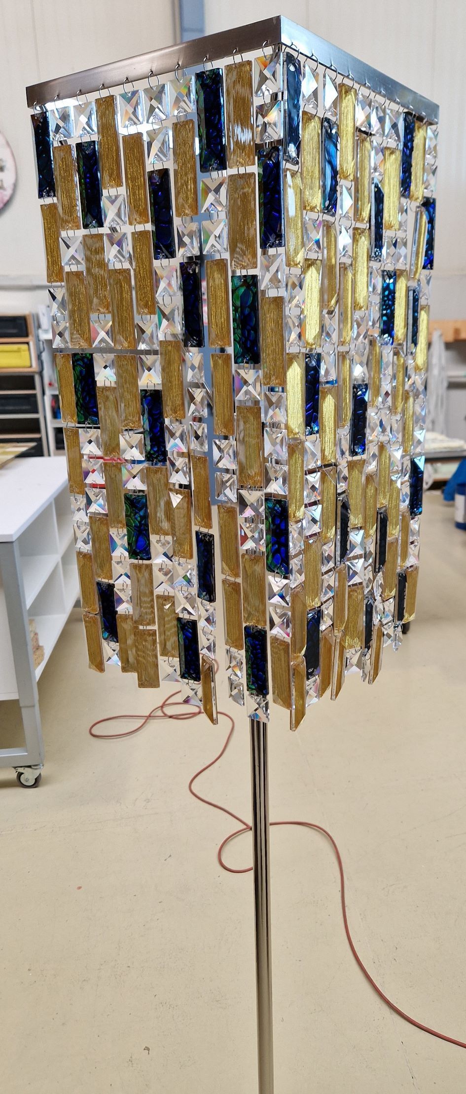 Ein quadratischer Lampenschirm aus rechteckigen Teilen in Gold, Silber und Blau hängt an einer Chromstange.