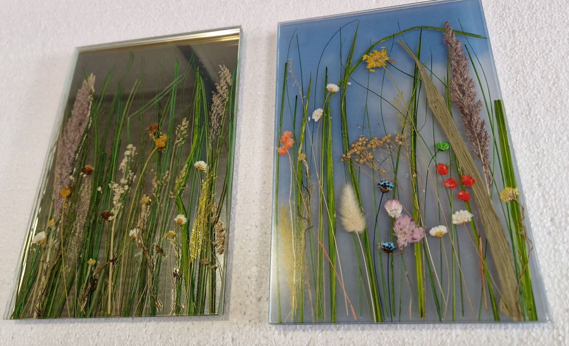 Zwei Glaskunstwerke: eines mit hohen Gräsern und gedeckten Farben, das andere mit blauem Hintergrund und bunten Blumen.