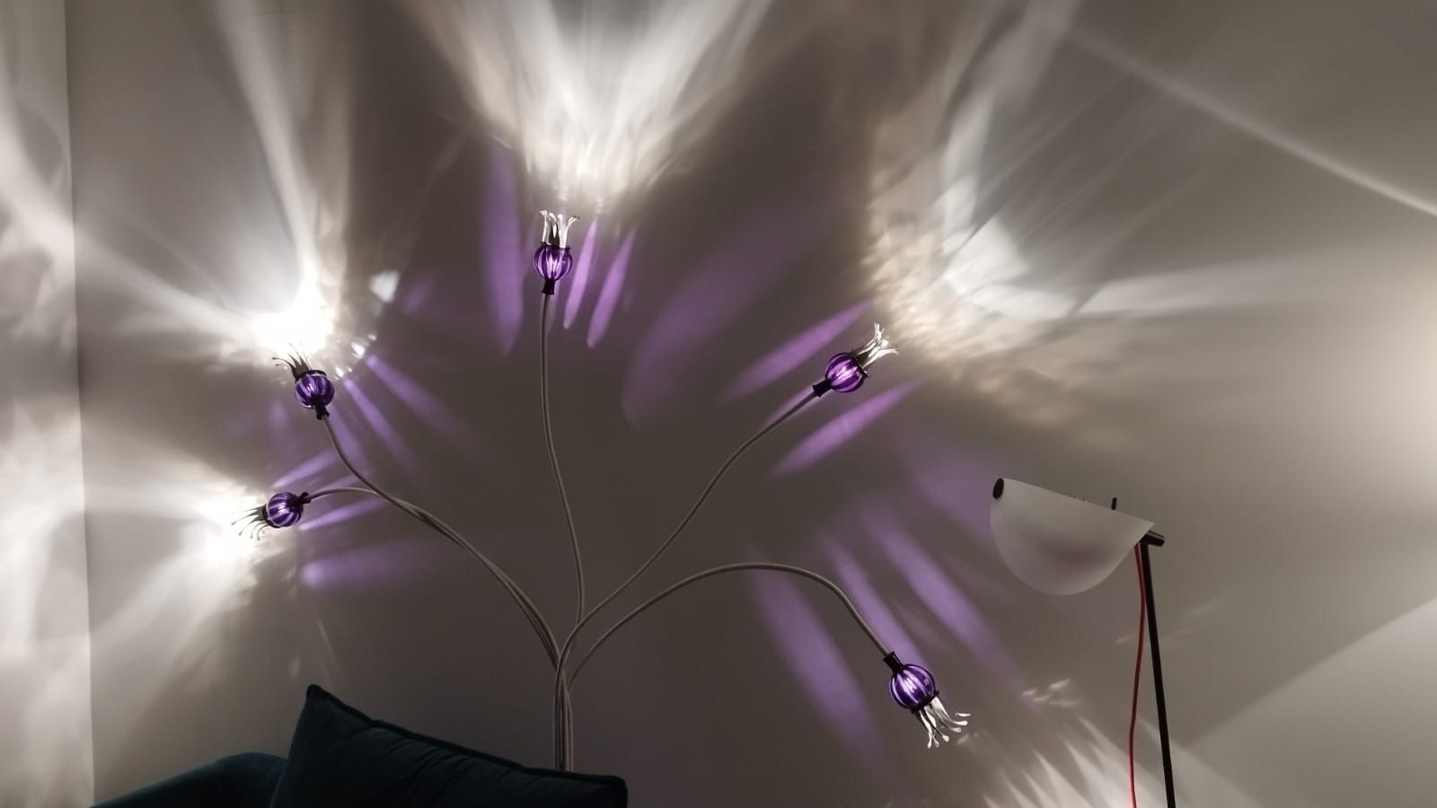 Lila beleuchtete dekorative Wandlampe mit sichtbarem Stromkabel, die Schatten auf eine weiße Wand wirft.