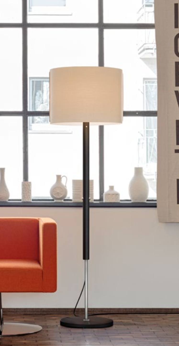 Stehlampe mit beigem Schirm und schwarz-silbernem Ständer, neben orangefarbenem Stuhl und Fenster mit weißen Vasen.