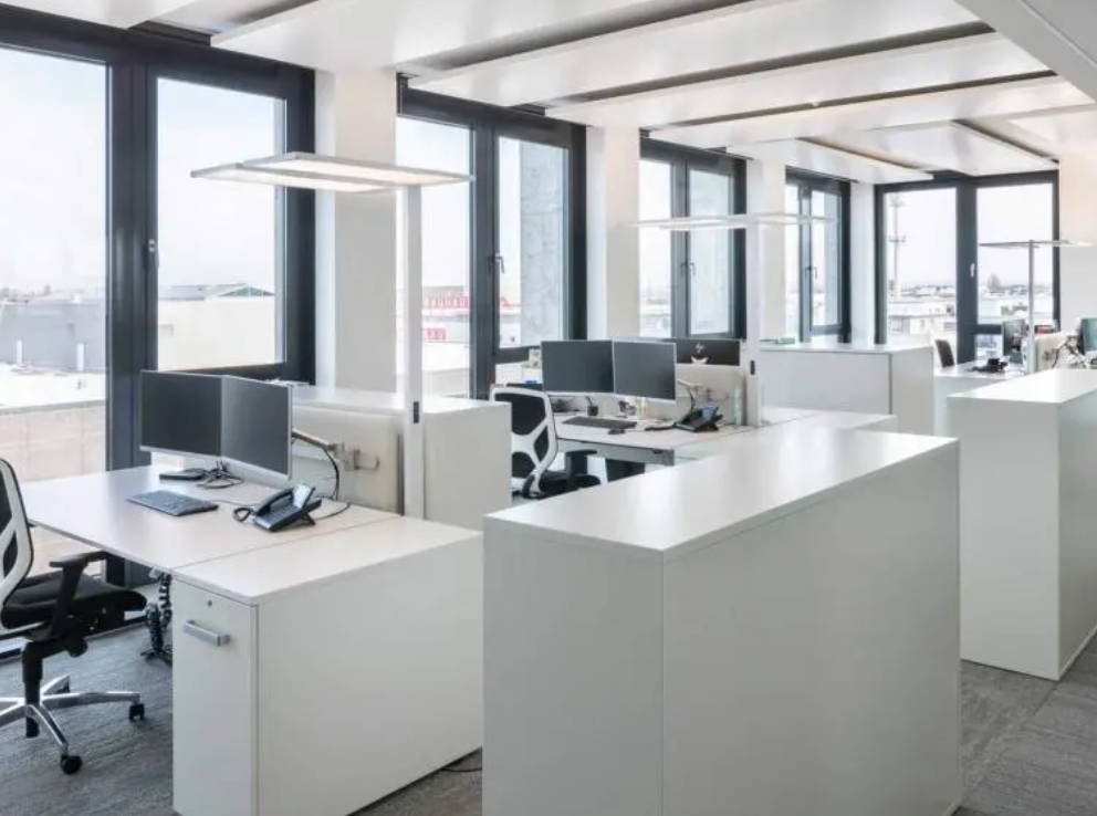 Helles, modernes Büro mit weißen Schreibtischen, Computern und schwarzen Stühlen in der Nähe großer Fenster.