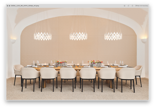 Langer Esstisch mit mehreren Stühlen, gedeckt für eine Mahlzeit. Darüber hängen drei moderne Kronleuchter.