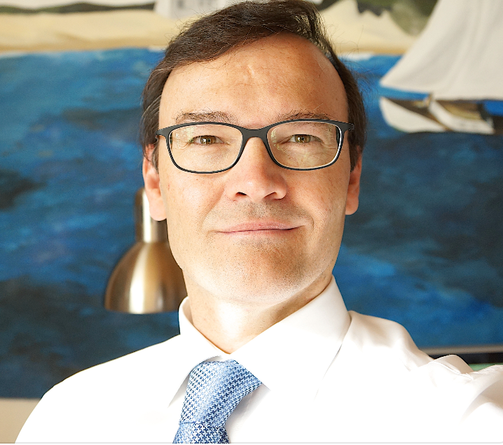 Mann mit Brille, lächelnd vor einem Meereslandschaftsgemälde, trägt ein weißes Hemd und eine blaue Krawatte.