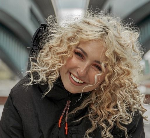 Frau mit blondem, lockigem Haar, lächelt breit und trägt einen schwarzen Mantel; im Freien mit einer Brücke im Hintergrund.