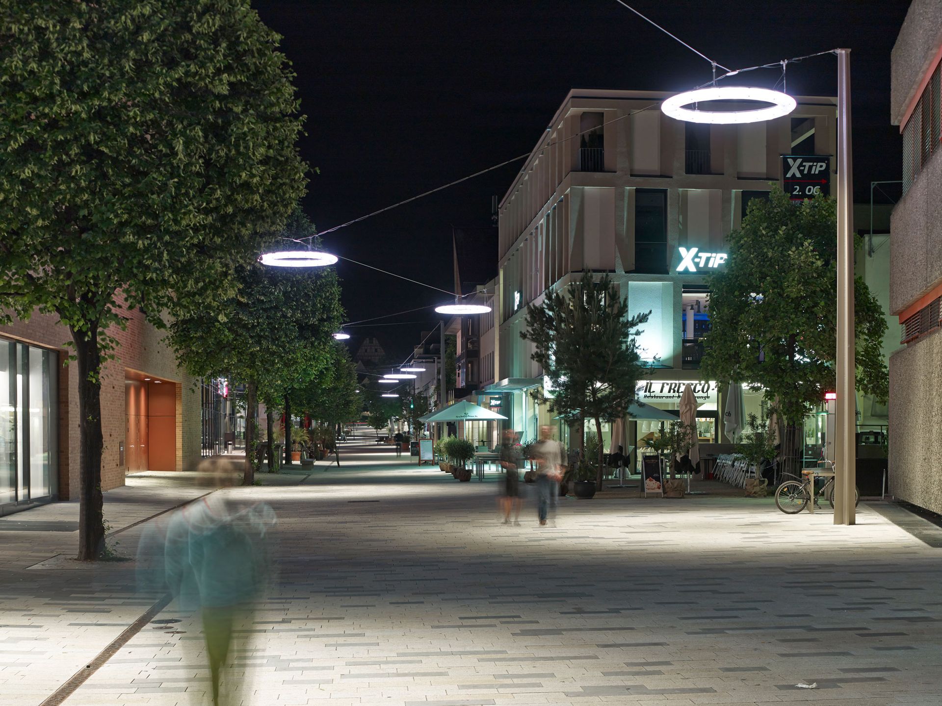Nächtliche Straßenszene mit Ringlichtern, Backsteingebäuden und verschwommenen Gestalten beim Gehen.