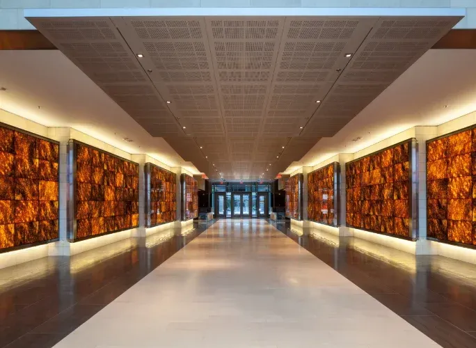 Langer Flur mit beleuchteten bernsteinfarbenen Paneelen an den Wänden. Darüber eine gemusterte Decke und ein reflektierender Boden.