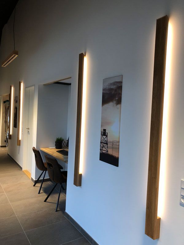 Wandleuchten mit Holzrahmen beleuchten einen weißen Flur mit Kunstwerken. Im Hintergrund ist ein Arbeitsbereich zu sehen.