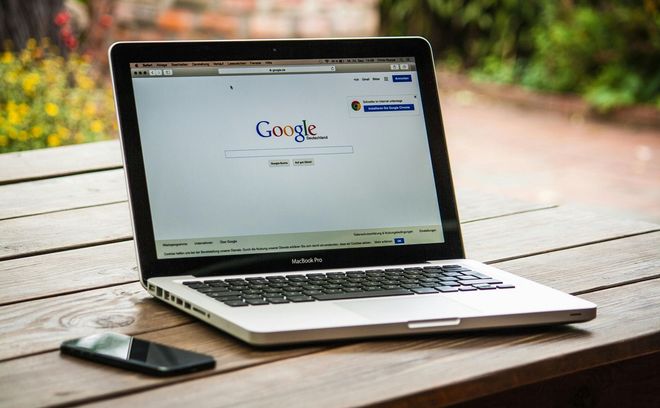 Laptop displaying Google search page, beside a smartphone on a wooden table outdoors.