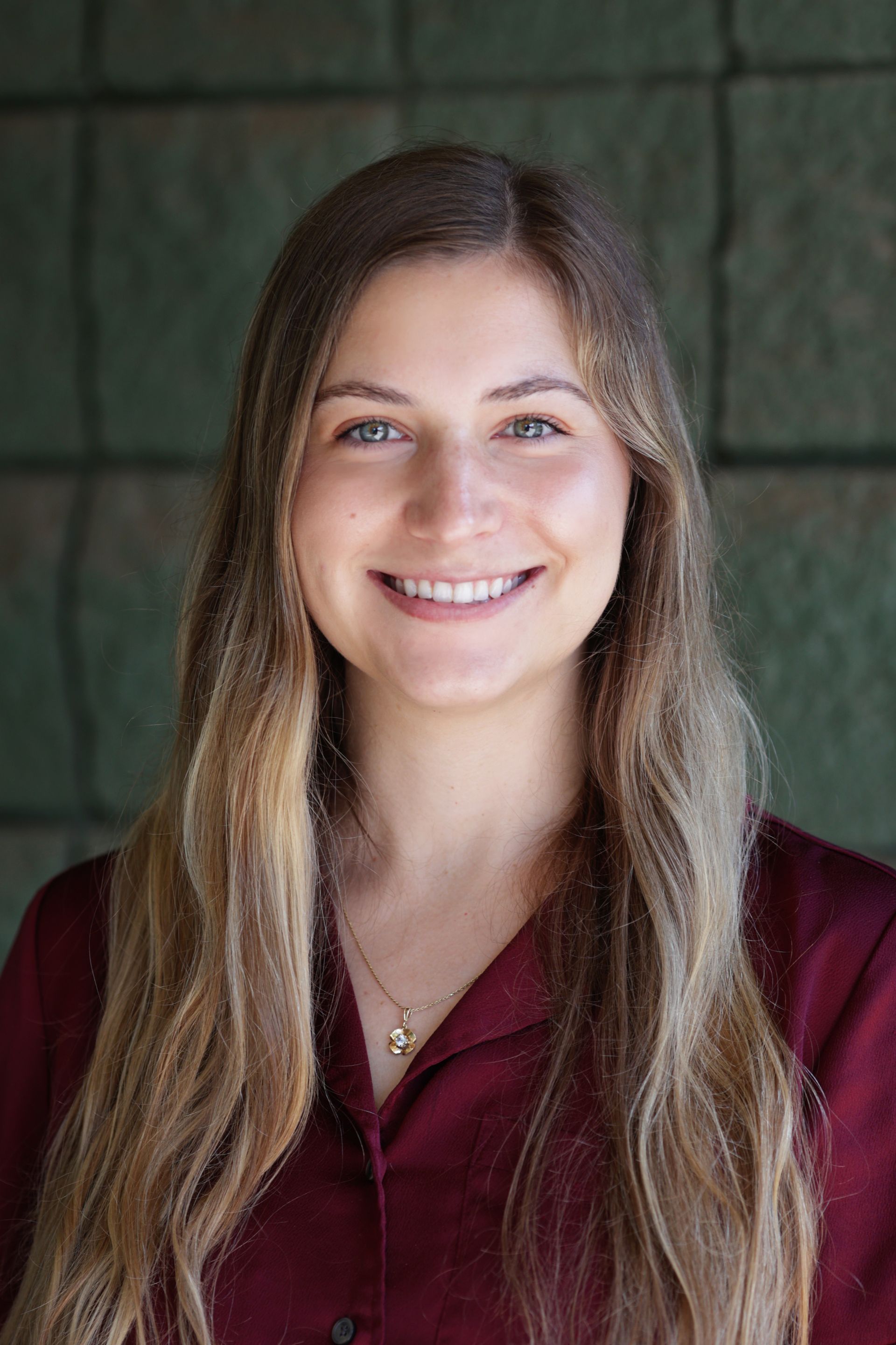 Woman with long wavy hair smiles at camera, wearing a maroon shirt, against a green brick background.