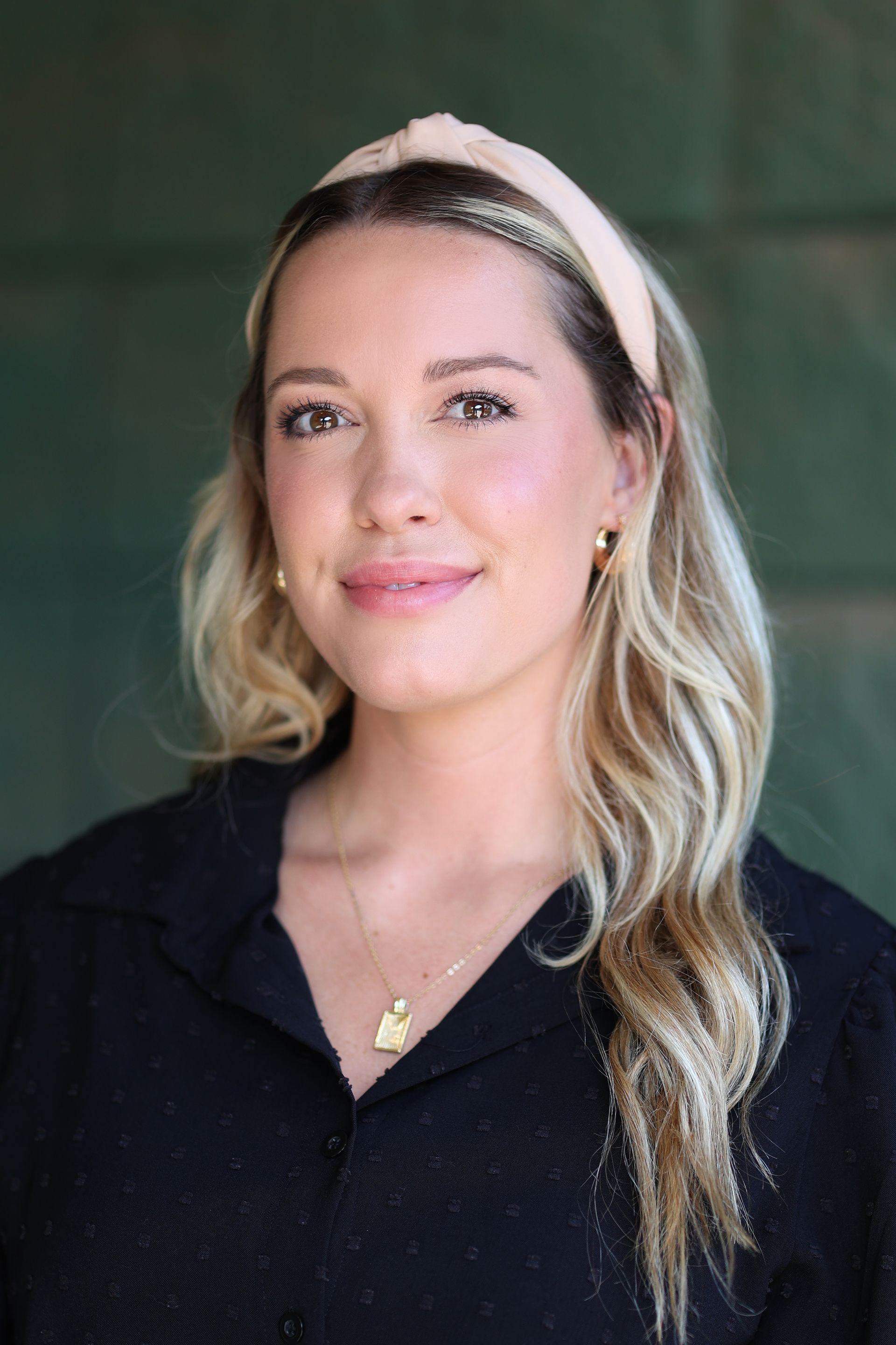 Woman with blonde hair and a headband smiles at the camera. She wears a black shirt, a necklace, and small earrings.