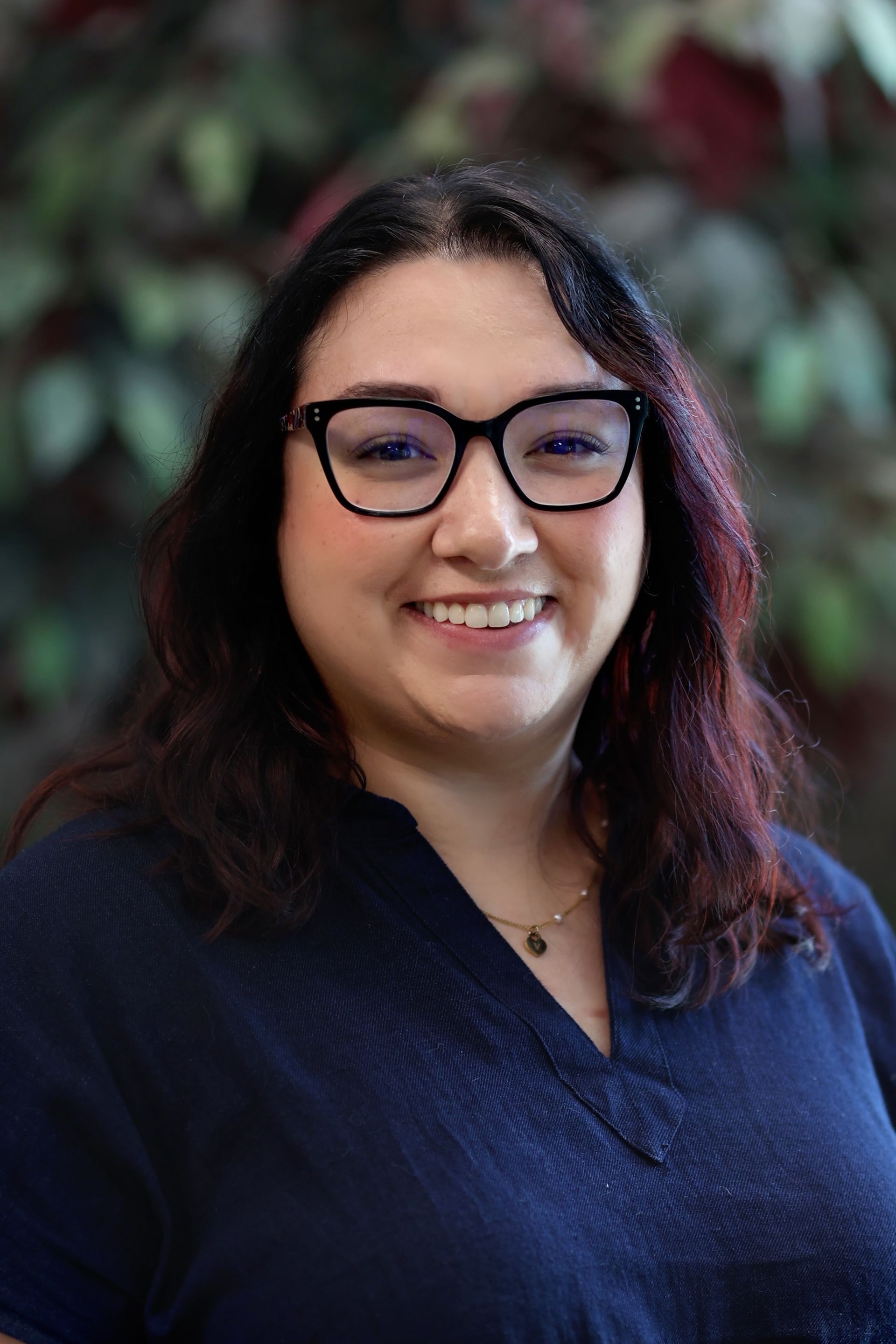 Woman with glasses smiling, wearing a dark blue shirt, in front of a blurred green and red background.