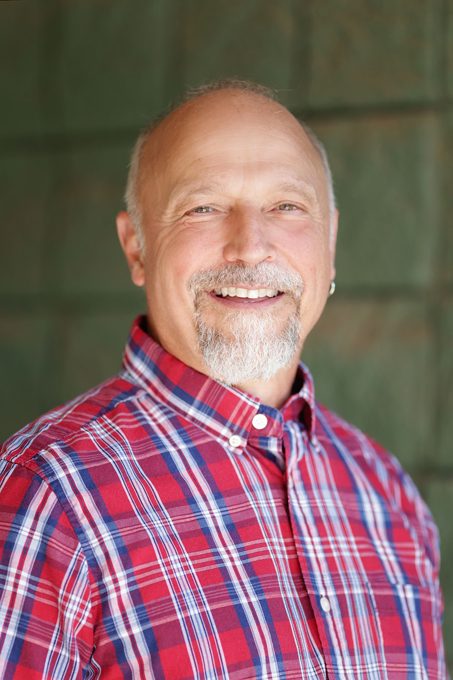 Smiling older man with a gray beard, wearing a red and blue plaid shirt, standing in front of a green wall.