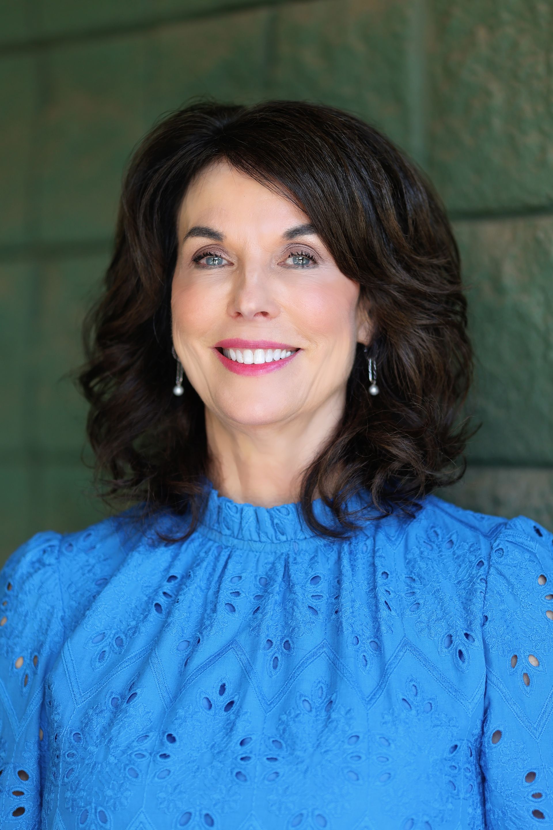 Woman with dark wavy hair smiles, wearing a blue eyelet blouse, posing in front of a green wall.