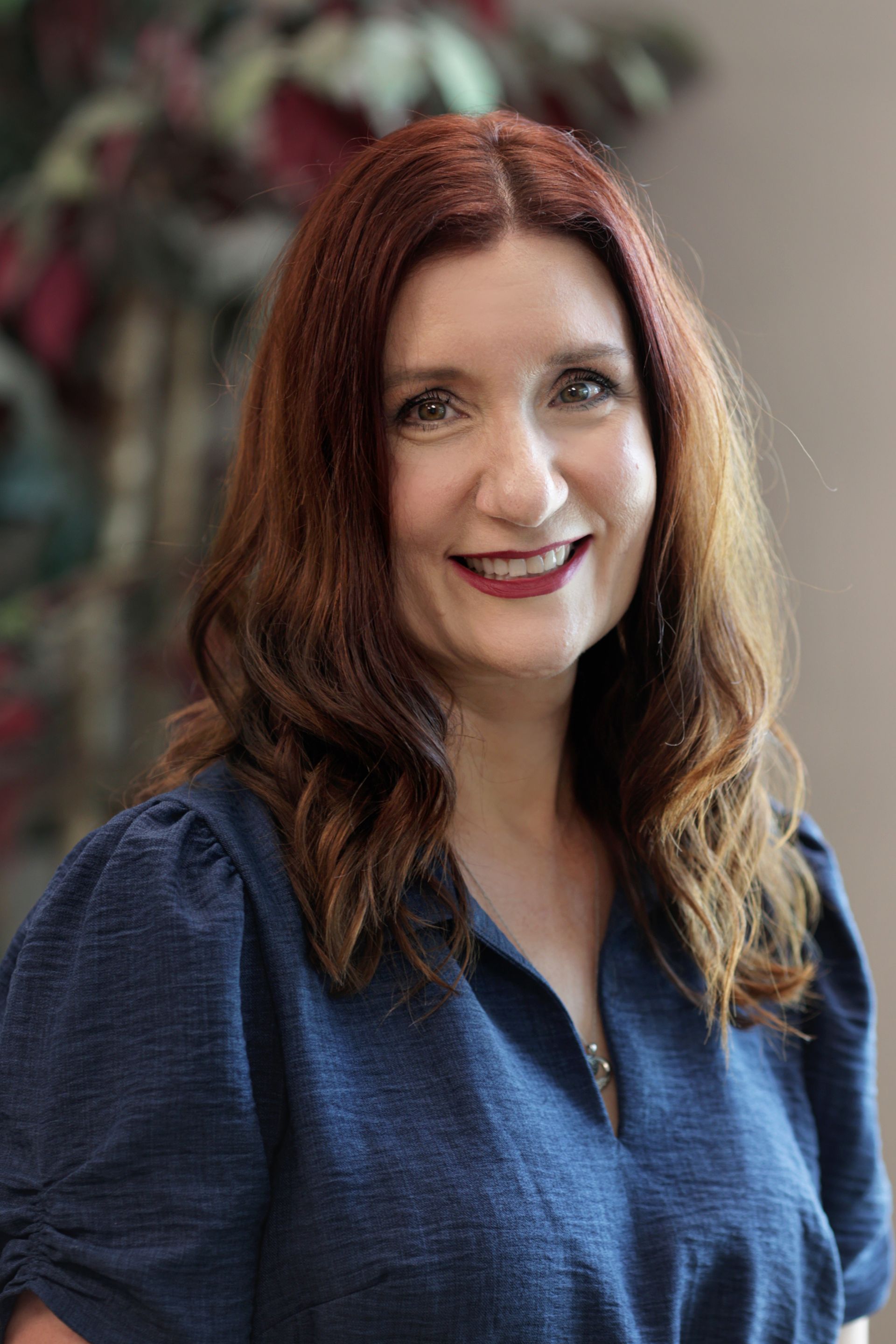 Woman with auburn hair smiles, wearing a blue textured top. Blurred background of foliage.