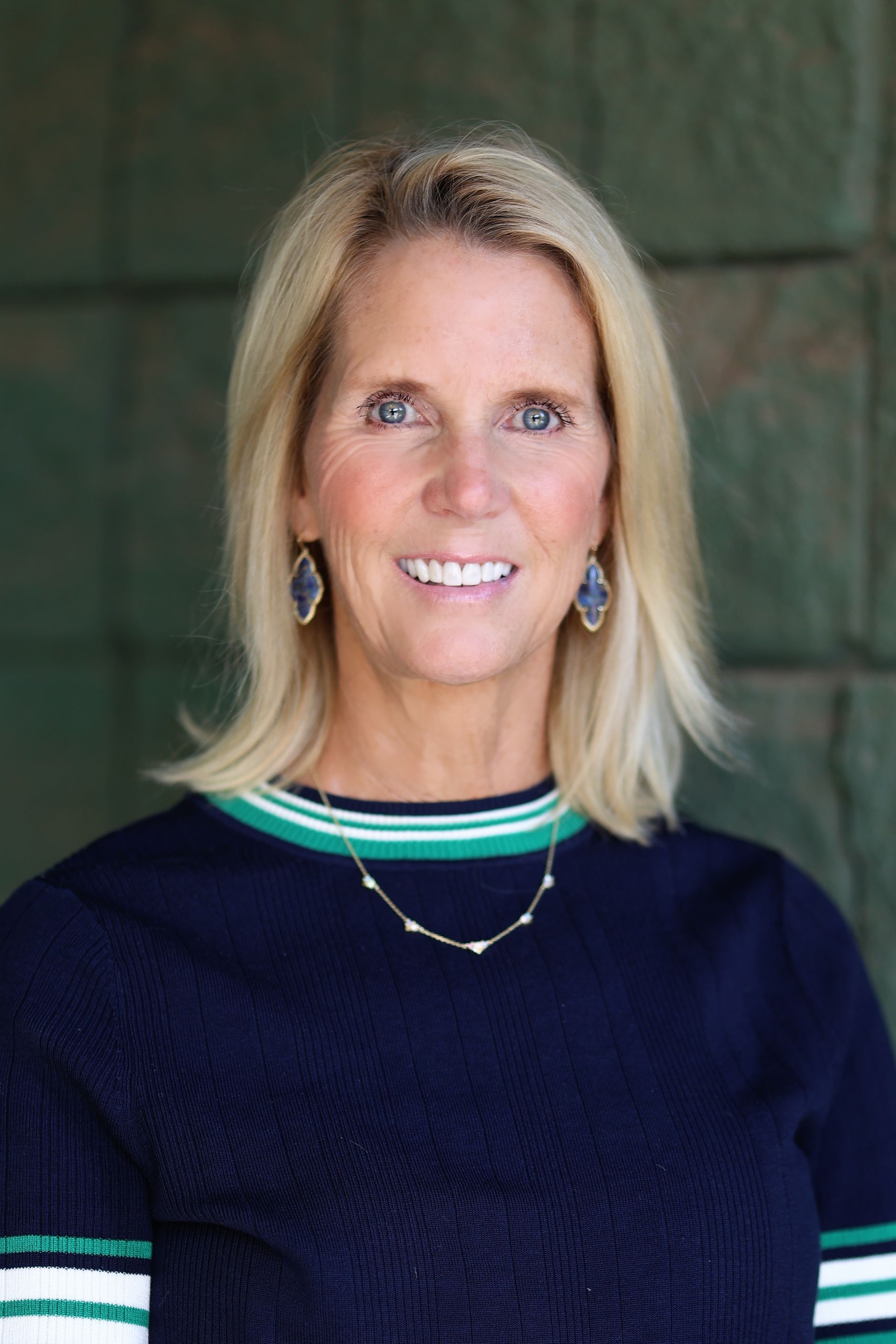 Blond woman with blue eyes smiles. She wears a navy sweater with green and white stripes and blue earrings.