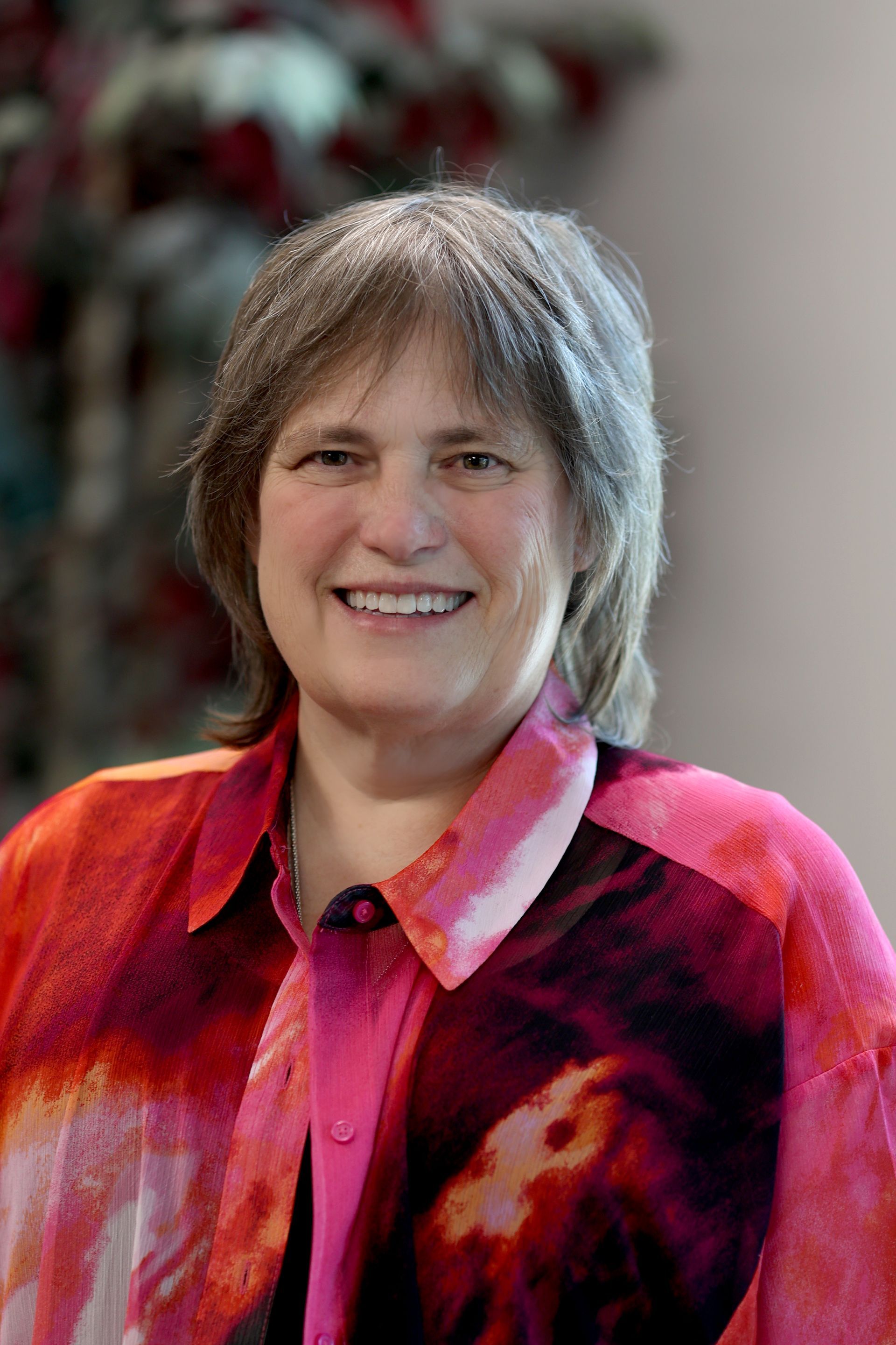 Woman with short gray hair, wearing a patterned red and pink button-down shirt, smiles. Background is blurry.