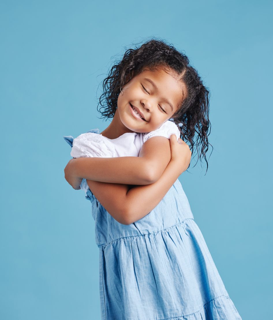 A little girl in a blue dress is hugging herself on a blue background.