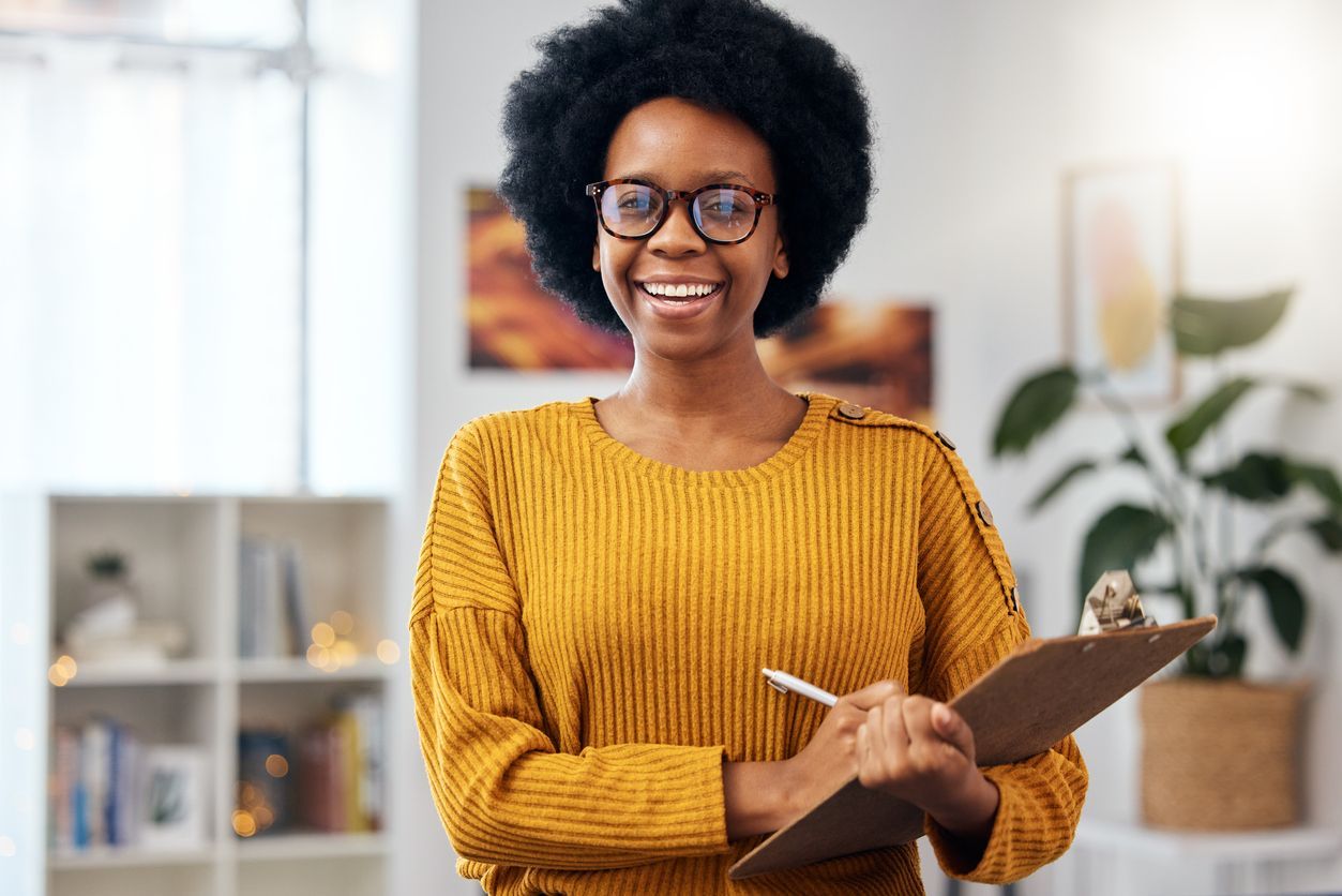 A woman in a yellow sweater is holding a clipboard and smiling.