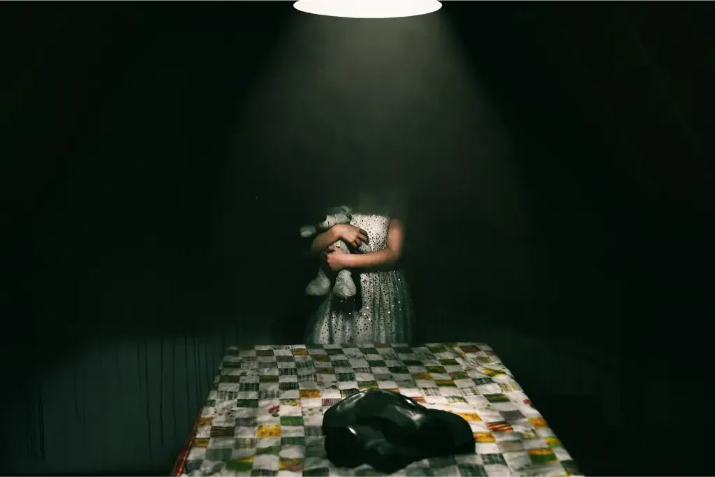 Een klein meisje zit aan een tafel in een donkere kamer en houdt een teddybeer vast.