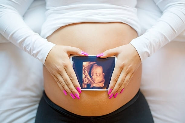 Close-up of a pregnant woman holding a 3D ultrasound photograph of a baby on the belly in bed.