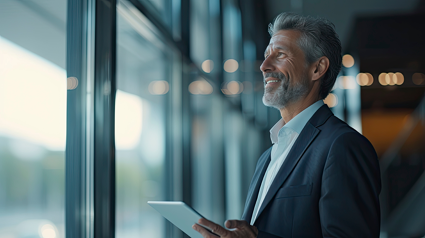 A man in a suit is holding a tablet and looking out of a window.