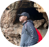 A man wearing a hat and a backpack is standing in front of a cave.