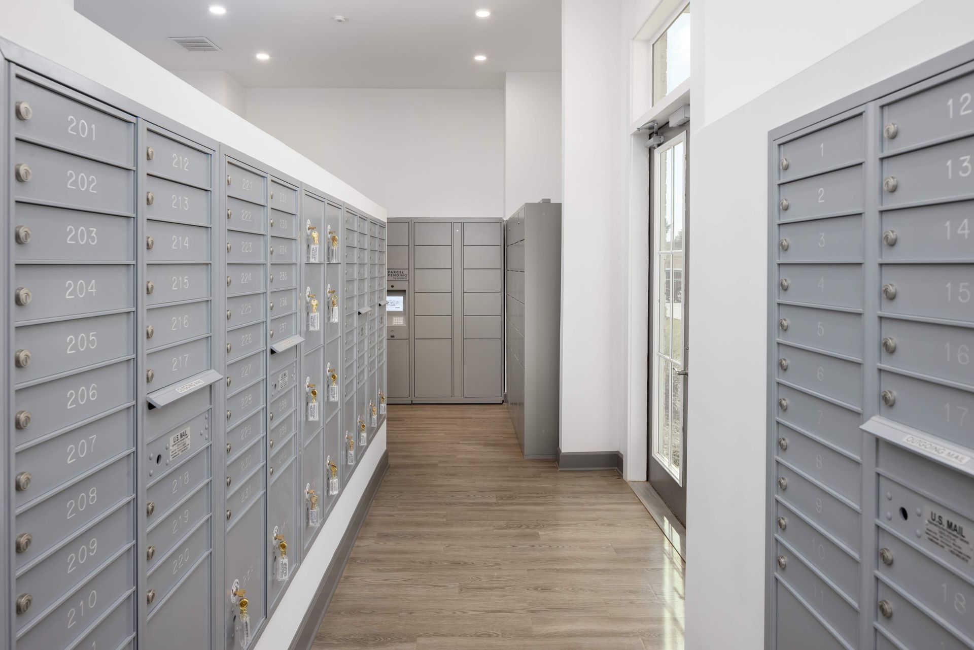 Mailboxes in a hallway, gray metal doors, wooden floor, lockers in background, door to outside.