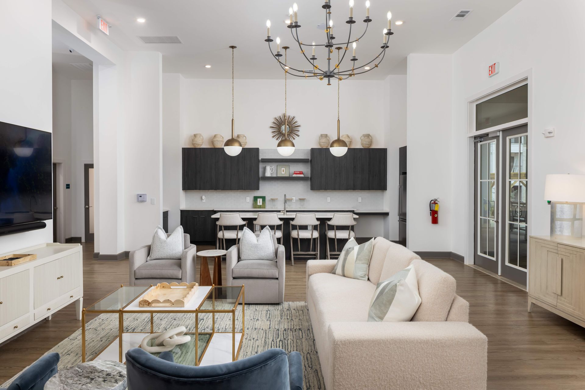 Lounge area with seating, a kitchen, and a chandelier. White walls, wood floor, neutral colors.