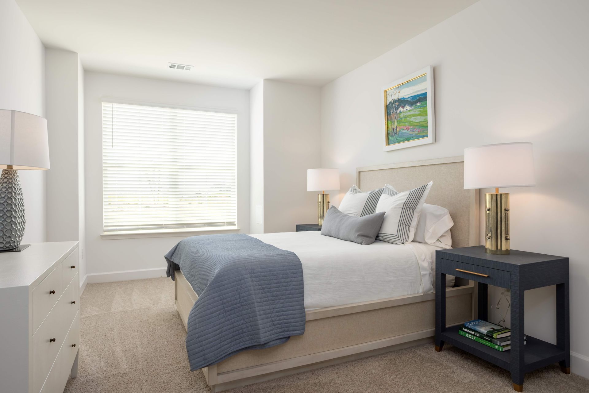 Bedroom with a bed, nightstand, dresser, and window. Neutral colors, blue blanket, and lamps.