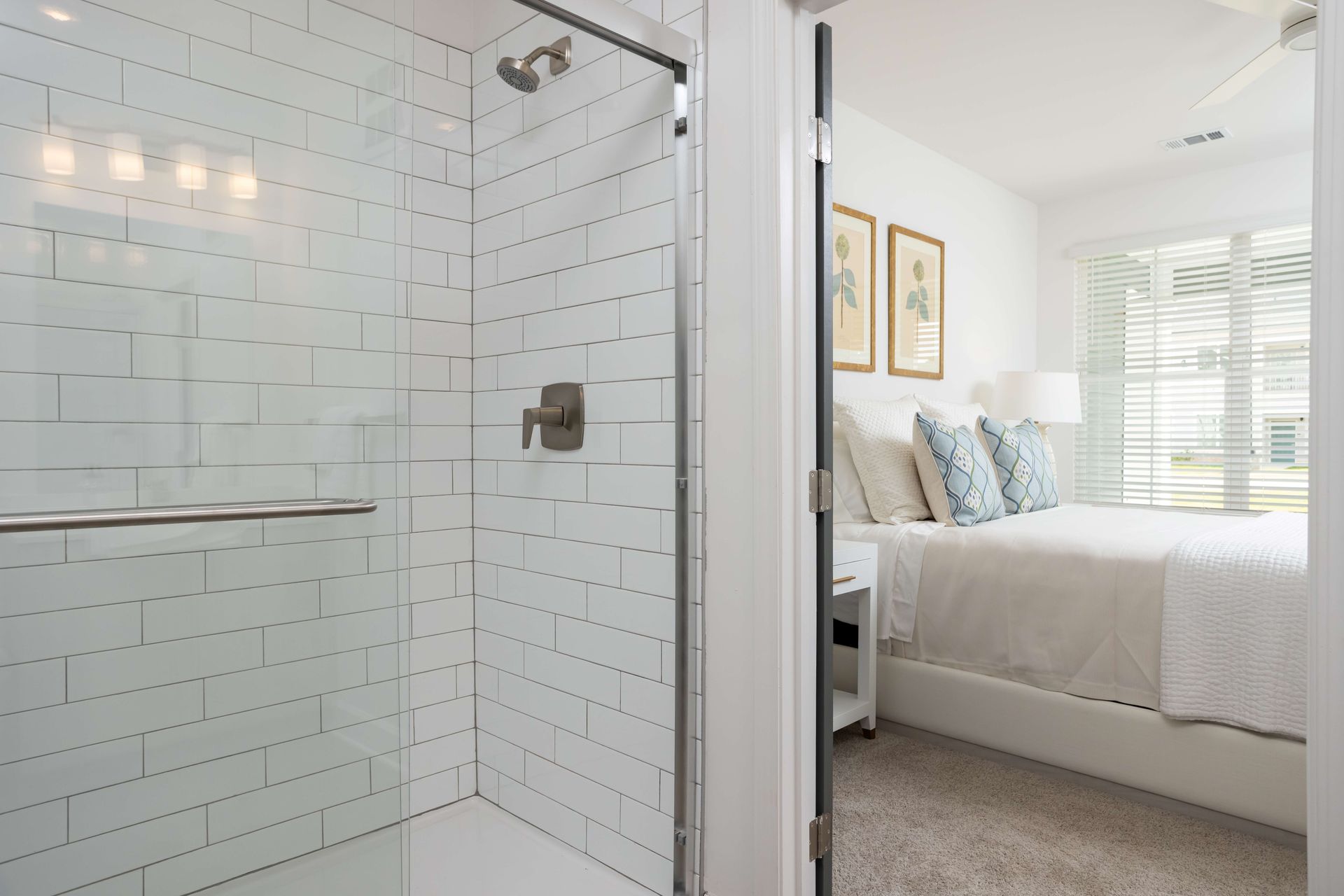 Shower with white tile, next to a bedroom with a bed and window with blinds.