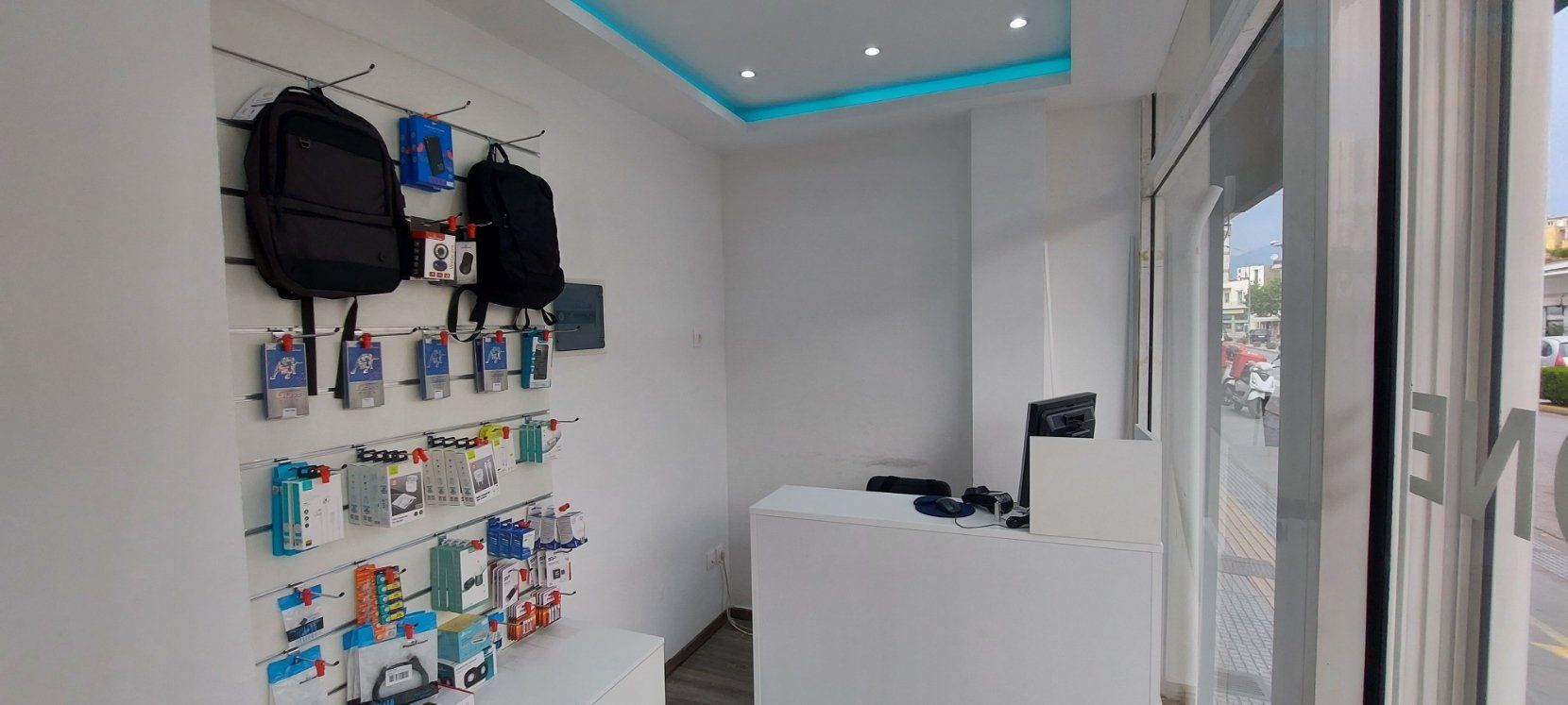 Εσωτερικό ενός μικρού καταστήματος αξεσουάρ τηλεφώνων με λευκούς τοίχους και βιτρίνες προϊόντων, φωτισμένο από ένα μπλε φωτιστικό οροφής.