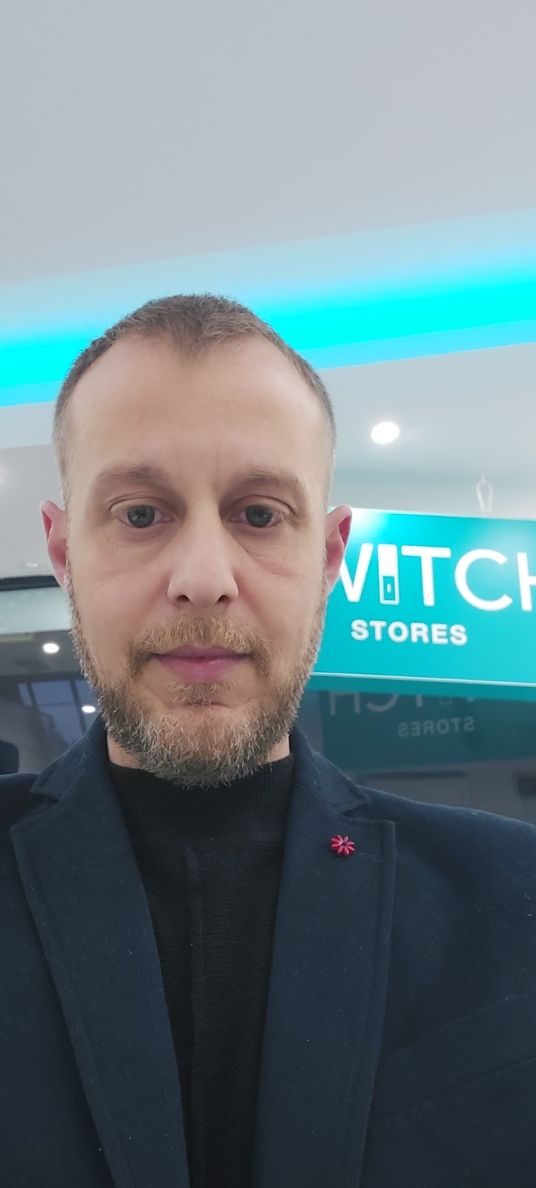 Άνδρας με σακάκι σε κατάστημα ρολογιών, στραμμένος προς την κάμερα. Έχει κοντή γενειάδα και ουδέτερη έκφραση.