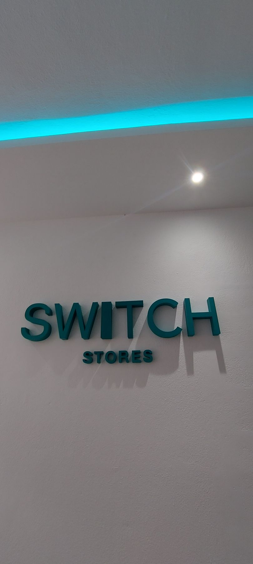 Πινακίδα για την ένδειξη «SWITCH STORES» σε γαλαζοπράσινο φόντο σε λευκό τοίχο, με μπλε φως από πάνω.