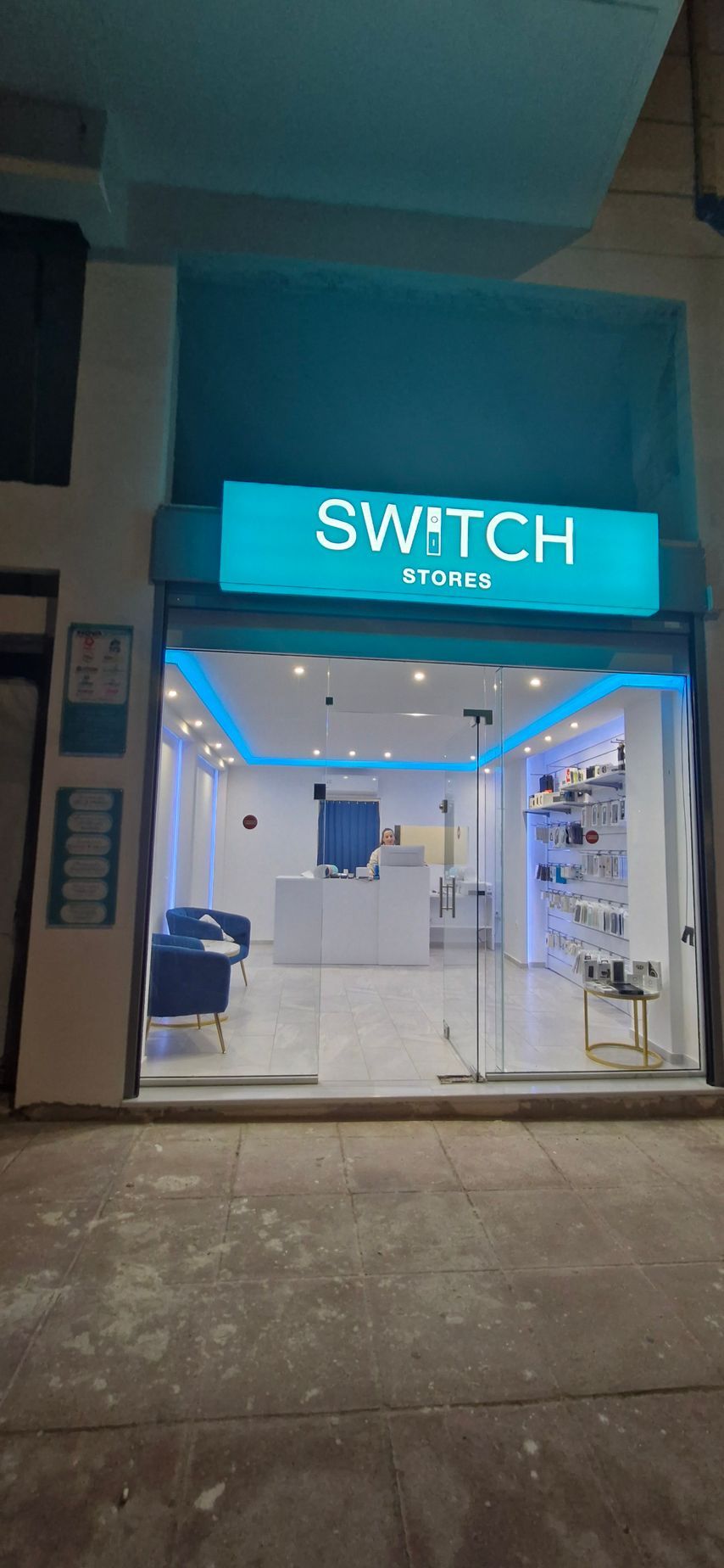 Εξωτερικό ενός καταστήματος που ονομάζεται Switch, με μια φωτεινή γαλαζοπράσινη πινακίδα και γυάλινη πρόσοψη, σε μια πόλη τη νύχτα.